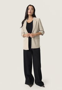 Blazer beige con maniche arrotolate, chiusura a un bottone e due tasche frontali, abbinato a un top nero aderente e pantaloni neri a gamba larga.