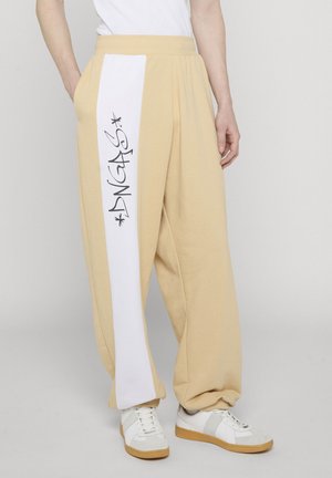 Bézs színű, bő szárú nadrág fehér függőleges csíkkal és fekete graffiti szöveggel a bal lábszáron, fehér sportcipővel és fehér felsővel párosítva.