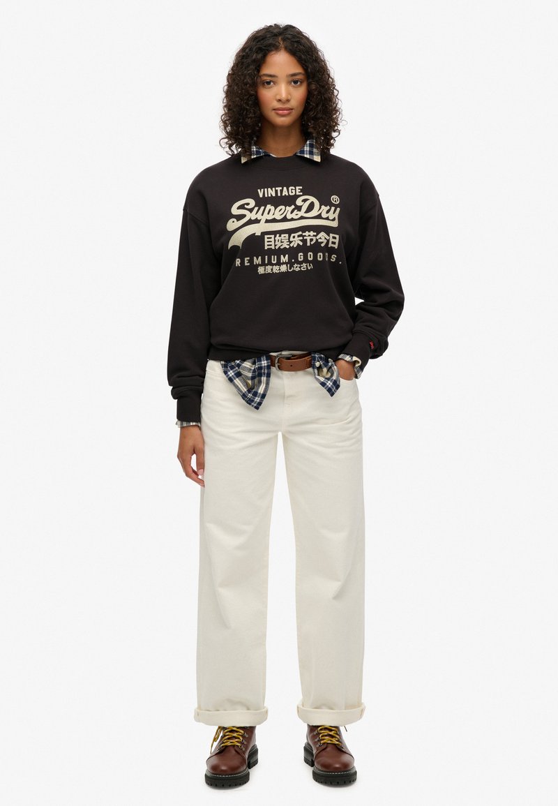 Felpa nera con logo "SuperDry", pantaloni larghi crema, camicia a quadri annodata in vita e stivali marroni con lacci e accentuazioni gialle.