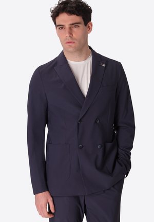 Uomo che indossa una giacca doppiopetto blu scuro e pantaloni abbinati, camicia bianca sotto, con una mano in tasca, guarda avanti.