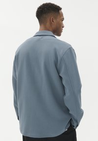 Man med en l�ös, långärmad blå skjorta sedd bakifrån mot en enkel vit bakgrund.