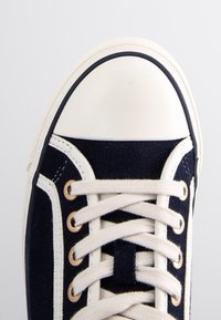 Baskets en toile bleu marine avec un embout en caoutchouc blanc et des œillets, dotées de lacets crème et de coutures contrastantes.