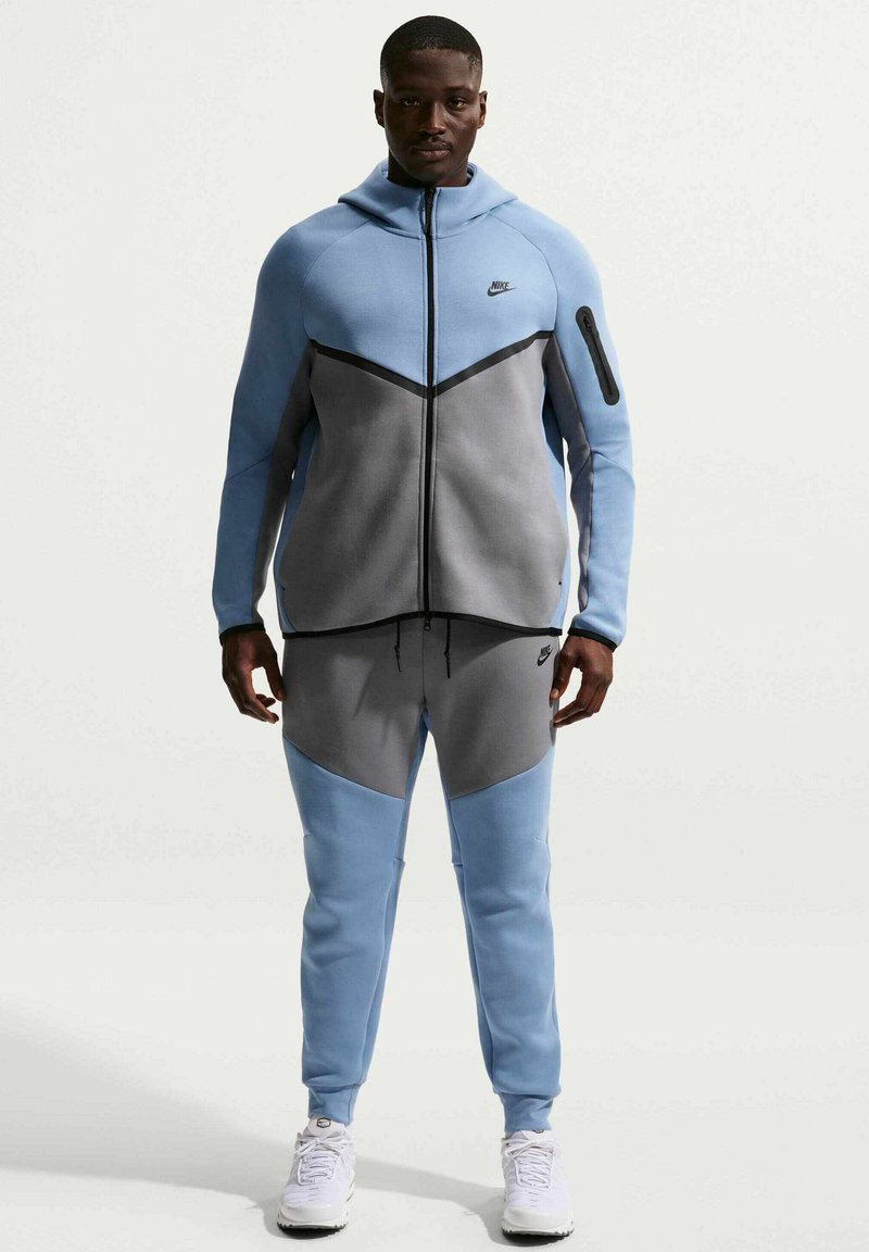 Tuta Nike con design a blocchi di colore azzurro e grigio, felpa con cappuccio e cerniera, e pantaloni abbinati, realizzata in tessuto morbido e testurizzato.