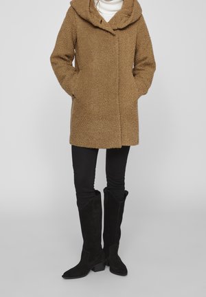 Tan textured coat with a hood, front button closure, and large pockets, paired with black slim-fitting pants and knee-high black suede boots.