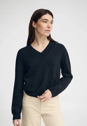 Kobieta o długich brązowych włosach nosi czarny sweter z dekoltem w serek i kremowe spodnie, patrząc w prawo na tle jednolitego jasnego tła.