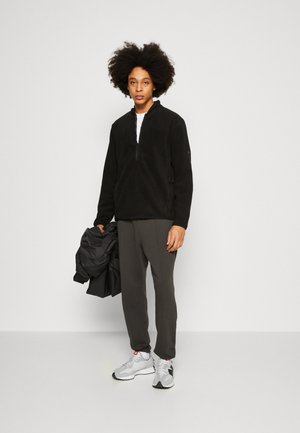 Μαύρο πουλόβερ από φλις με μισό φερμουάρ, συνδυασμένο με σκούρο γκρι παντελόνι και γκρι αθλητικά παπούτσια. Το σύνολο διαθέτει λεία υφή και αθλητική σχεδίαση.