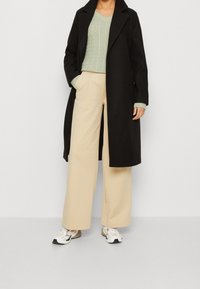 Schwarzer langer Mantel, hellgrüner Kabelstrick-Pullover, beigefarbene weite Hose und weiße Sneakers. Minimalistisches Design mit strukturierten Materialien.