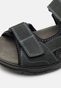 Tumman vihreä nahkasandaali, jossa on säädettävät Velcro-nauhat, kontrastivärinen harmaa ommel ja teksturoitu musta pohja pidon takaamiseksi. Pehmeä pohja näkyvissä.