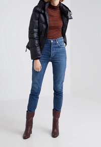 Kvinde iført sort dunjakke, rust- og sortstribet langærmet top, højtaljede blå jeans og mørkebrune støvler med hæl.