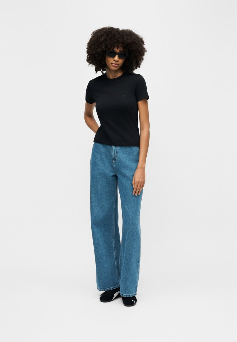 Woman wearing black sunglasses, black fitted T-shirt, wide-leg blue jeans, and black sneakers, standing with one hand behind her back.