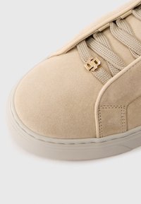 Bež antilop tenisica s zaobljenim vrhom, teksturiranim gornjim dijelom, ravnim vezicama i metalnim zlatnim detaljem pokraj vezica. Bijeli gumeni potplat.