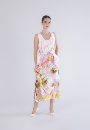 Donna con un vestito di seta rosa senza maniche con un motivo floreale multicolore e tacchi alti bianchi, in piedi davanti a uno sfondo chiaro uniforme.