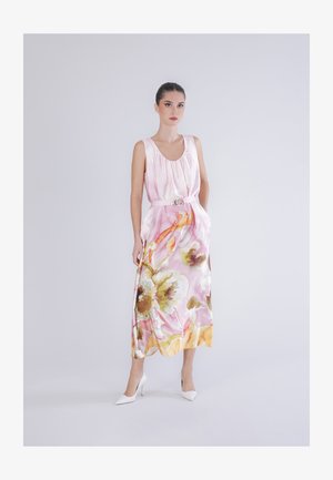 Vrouw in een mouwloze roze zijden jurk met een multicolor bloemenpatroon en witte hoge hakken, staand tegen een effen lichte achtergrond.