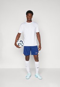 Biela športová tričko, námornícke šortky s oranžovými akcentmi, biele ponožky po kolená, svetlomodré futbalové topánky a voľné vzorované futbalové lopty.