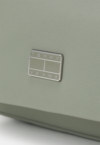 Borsa in pelle verde con superficie texturizzata. Presenta una piastra logo rettangolare metallica incisa con "TOMMY" e "JEANS" nella parte inferiore.