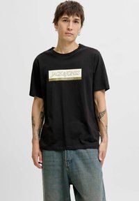 Sort bomulds T-shirt med et fremtrædende gyldent rektangulært logo med teksten "JACK & JONES". Korte ærmer og afslappet pasform, parret med lyseblå jeans.