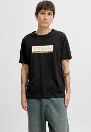Camiseta negra de algodón con un prominente logo rectangular dorado con el texto "JACK & JONES". Mangas cortas y ajuste relajado, combinada con jeans azules claros.