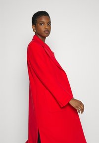 Roter Wollmischungsmantel mit einem langen, lockeren Schnitt, breiten Revers und seitlichen Taschen, der eine glatte Textur und einen seitlichen Schlitz für Bewegungsfreiheit bietet.