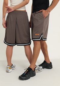 Shorts de sport en mesh gris avec des bordures noir et blanc, mettant en avant un accent logo orange. Portés par deux personnes de la taille vers le bas, montrant différentes chaussures.