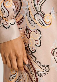 Fließendes Satinkleid mit Paisleymuster, das in Beigetönen, Schwarz und multicolor Akzenten gehalten ist. Nahaufnahme von Detail des Ärmels und der Hand.