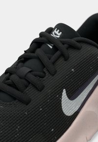 Fekete sportcipő texturált hálós felsőrészsel, amelyen egy jellegzetes fehér Nike kampó található, fekete fűzőkkel és rózsaszín gumitalppal.