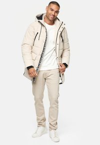 Beige polstret jakke med hætte, sorte detaljer og to forlommer. Båret over en hvid sweater og lyse beige bukser med hvide sneakers.