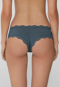 Krajková tanga v tmavě modré barvě s hladkým tělem a vlnitými okraji, navržená pro pohodlí a těsné přilnutí.