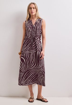 Kvinde iført en ærmeløs kjole til anklerne med et brunt og beige abstrakt zebra mønster og sorte sandaler, stående op ad en ensfarvet væg.