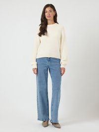 Kobieta stoi w kremowym swetrze, jasnoniebieskich prostych dżinsach i beżowych szpilkach na tle szarego tła.