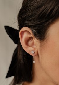 Pendientes de plata con un diseño floral que presentan un aro con rhinestones y un colgante de cadena que termina en un acento en forma de pera. Cinta negra en el cabello.
