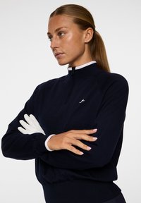 Merinopullove at half-zip meren oikealla läpällä ja helmassa. Siinä on valkoinen logo rinnassa ja se korostaa tasaista, teksturoidua kangasta.