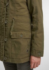 Olivově zelená plátěná bunda s knoflíkovým zapínáním, nastavitelným stahováním v pase, náprsní kapsou a detaily s knoflíky na manžetách.
