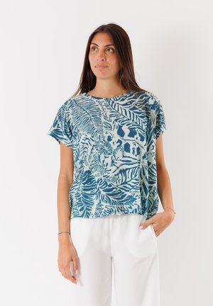 Top a maniche corte con un motivo di foglie blu e turchese, caratterizzato da una vestibilità morbida e un tessuto leggero. Indossato con pantaloni bianchi.