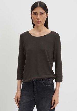 Vrouw met lang donker haar, gekleed in een donker olijfgroen longsleeve en zwarte jeans, staand tegen een effen lichte achtergrond.