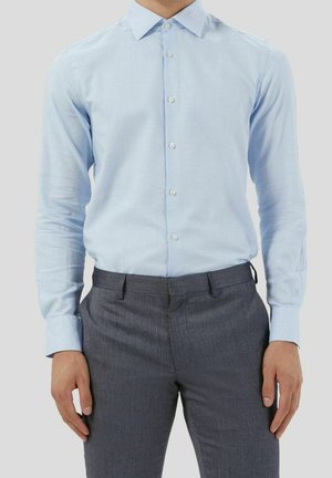 Homme portant une chemise bleu clair à manches longues boutonnée, rentrée dans un pantalon de costume gris, debout devant un fond clair uni.