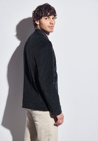 Černá kordurová bunda s přiléhavým střihem, zapínáním na jeden knoflík a jemným vertikálním žebrováním. Bílý pozadí zdůrazňuje detaily oděvu.