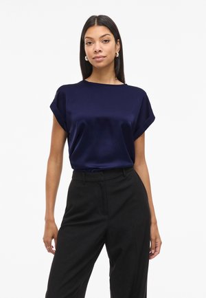 Nő tengerészkék rövid ujjú szatén blúzban és magas derekú fekete nadrágban, egyszínű fehér háttér előtt állva.