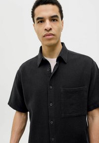 Chemise noire à manches courtes avec un tissu texturé, une poche poitrine et un col classique. Les boutons sont ronds et foncés.