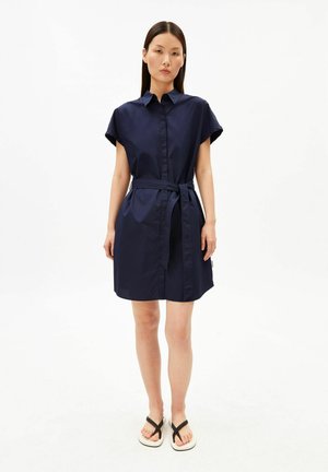 Femme portant une robe-chemise bleu marine à manches courtes avec une ceinture nouée à la taille et des sandales noires et blanches, debout devant un fond blanc uni.