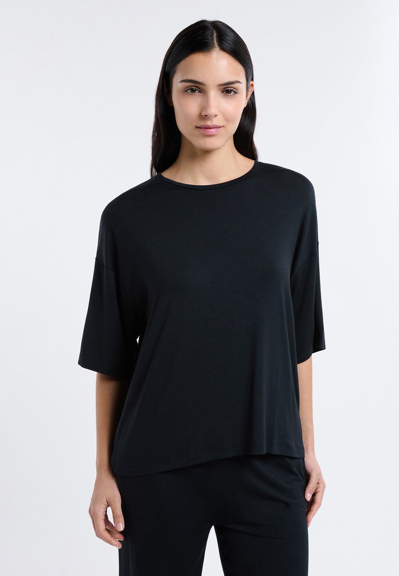 Femme aux longs cheveux noirs portant un haut noir ample à manches courtes et un pantalon noir, debout devant un fond clair uni.
