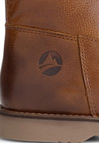 Bruine leren boot met een gladde textuur, subtiele stiksels en een rond logo met een bergontwerp aan de buitenkant. De zool is in een lichte kleur.