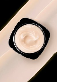 Crème dans un pot noir rond, avec une texture lisse et beige clair. Placée sur un fond pâle et fluide avec un bord sombre.