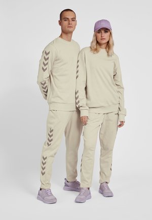 Due modelli indossano tute beige coordinati con strisce a chevron, sneakers lavanda, e un modello indossa un berretto lavanda.