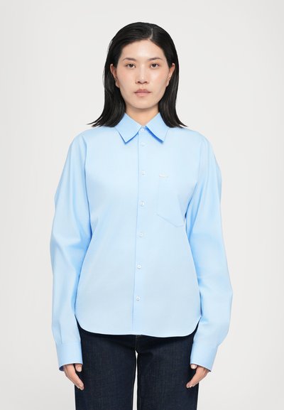 Helleblaue Button-Down-Bluse mit klassischem Kragen, langen Ärmeln, einer Brusttasche und einer glatten Stofftextur. Kombiniert mit dunklen Jeans.