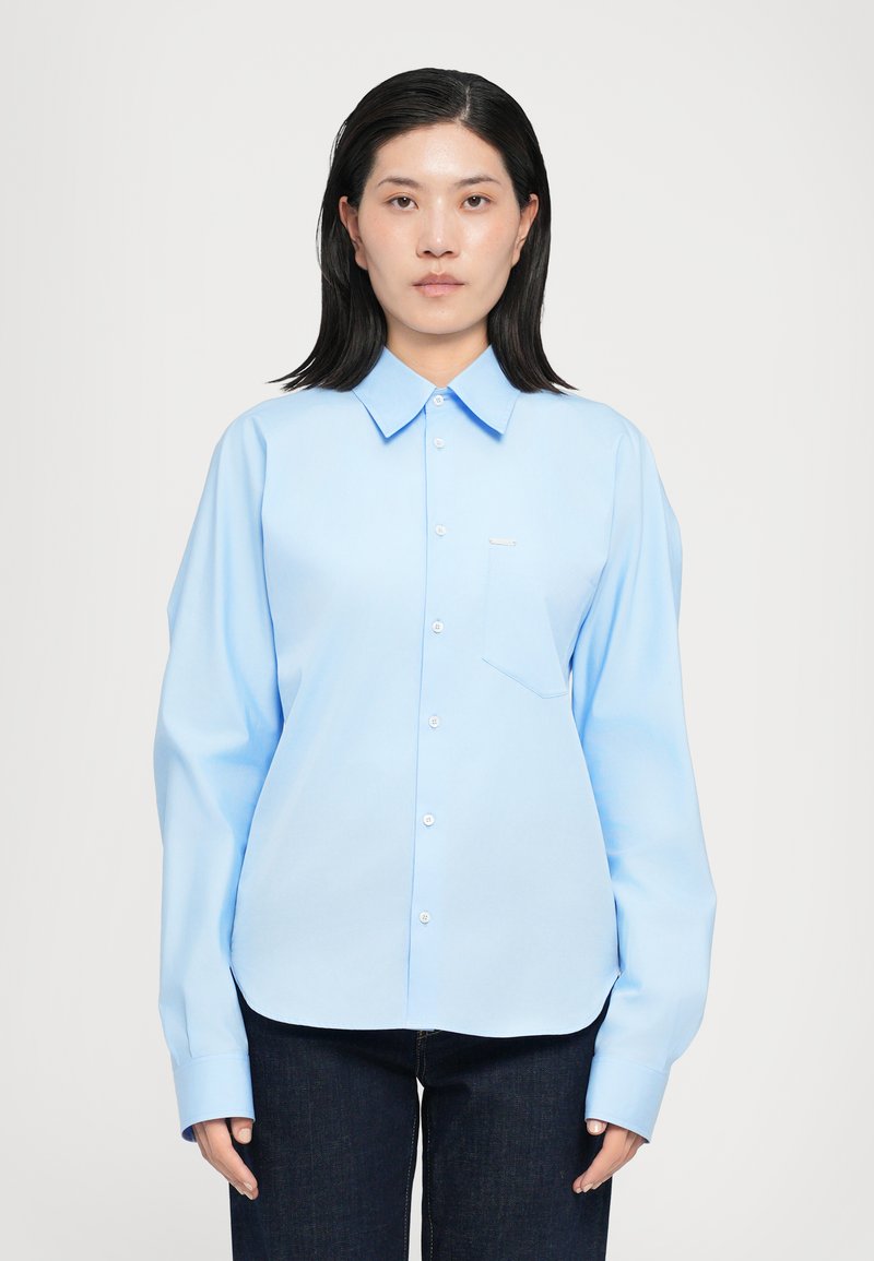Light blue button-up shirt with a classic collar, long sleeves, a chest pocket, and a smooth fabric texture. Paired with dark jeans.