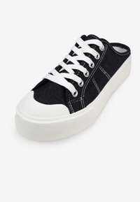 Zapatillas de lona negras con suelas de goma blanca, puntera redonda y cordones blancos. Presentan ojales de plata y acentos de costura.