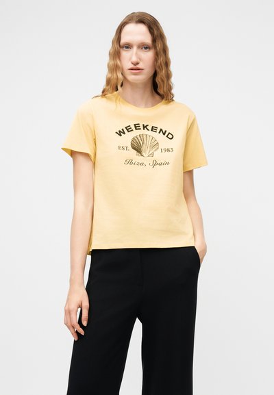 Femme aux cheveux bruns clairs ondulés portant un T-shirt jaune avec l'inscription "WEEKEND EST. 1983 Ibiza, Espagne" et un motif de coquillage, associé à un pantalon noir.