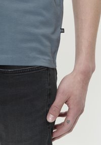 Camiseta de algodón gris con escote redondo, mangas cortas y una etiqueta de lado. Combinada con jeans de mezclilla negros, con un primer plano de una mano.