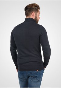 Donkergrijs gebreid sweater met hoge kraag, voorzien van elleboogpatches en een geribbelde zoom. Gecombineerd met blauwe spijkerbroek, die een getailleerd silhouet toont.
