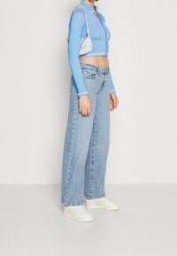 Kobieta w niebieskiej krótkej bluzie na zamek, jasnoniebieskich szerokich dżinsach, białych trampkach i małej wzorzystej torbie na ramię.